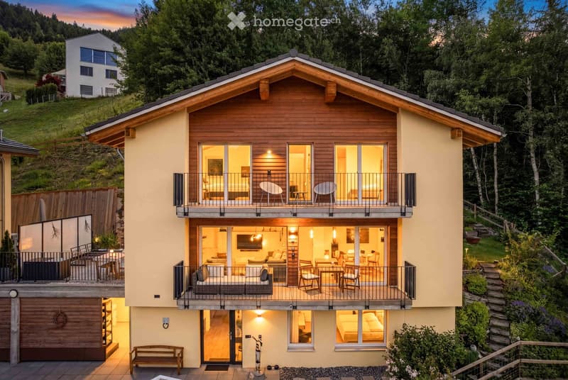Modern two-story house with wooden accents, large windows, balconies, and a garage. The house is surrounded by greenery and trees.