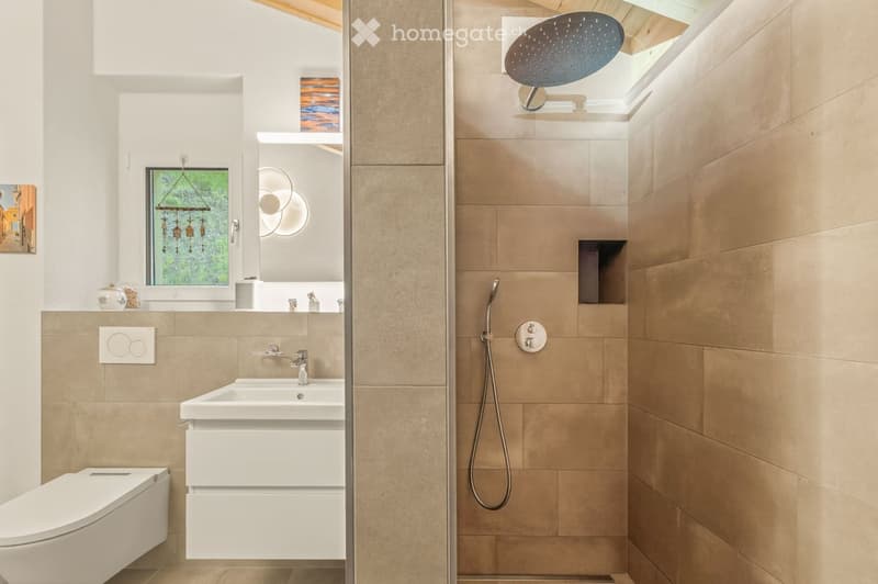 Modern bathroom with beige tiles, white sink, toilet, shower area with handheld showerhead, and a large window with decorative hanging ornament.