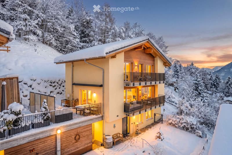 A modern, two-story house with a snow-covered roof and surrounding snowy landscape. The house features large windows, balconies, and a well-lit interior visible through the glass. The exterior includes a wooden garage and a small outdoor seating area.