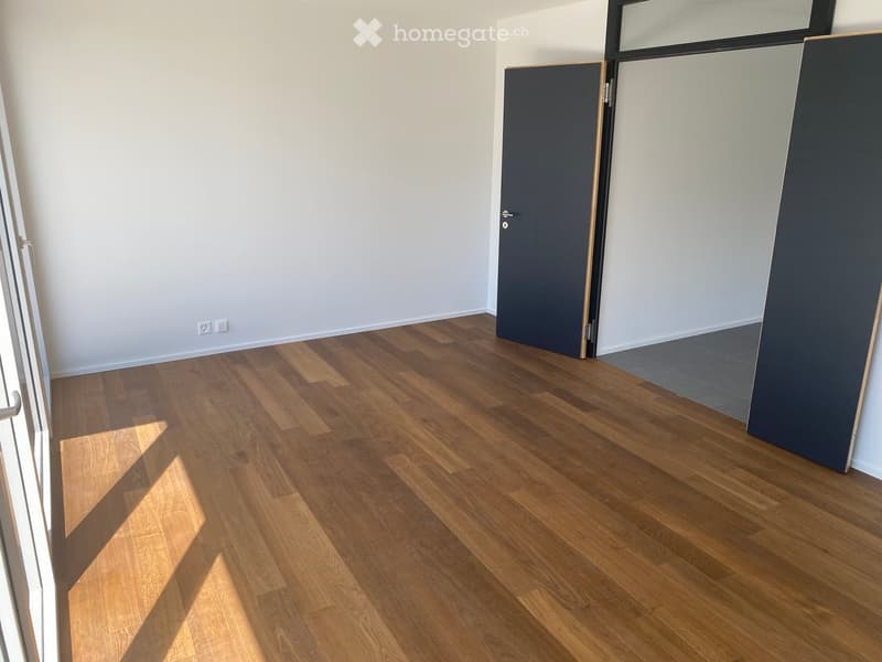 Empty room with light-colored walls and wooden flooring, featuring a closed door and a partially visible window frame.