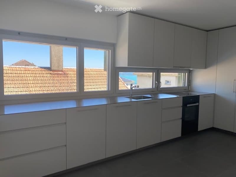 Modern, minimalist kitchen with white cabinets, a central island, and large windows offering a view of the roof and sea.