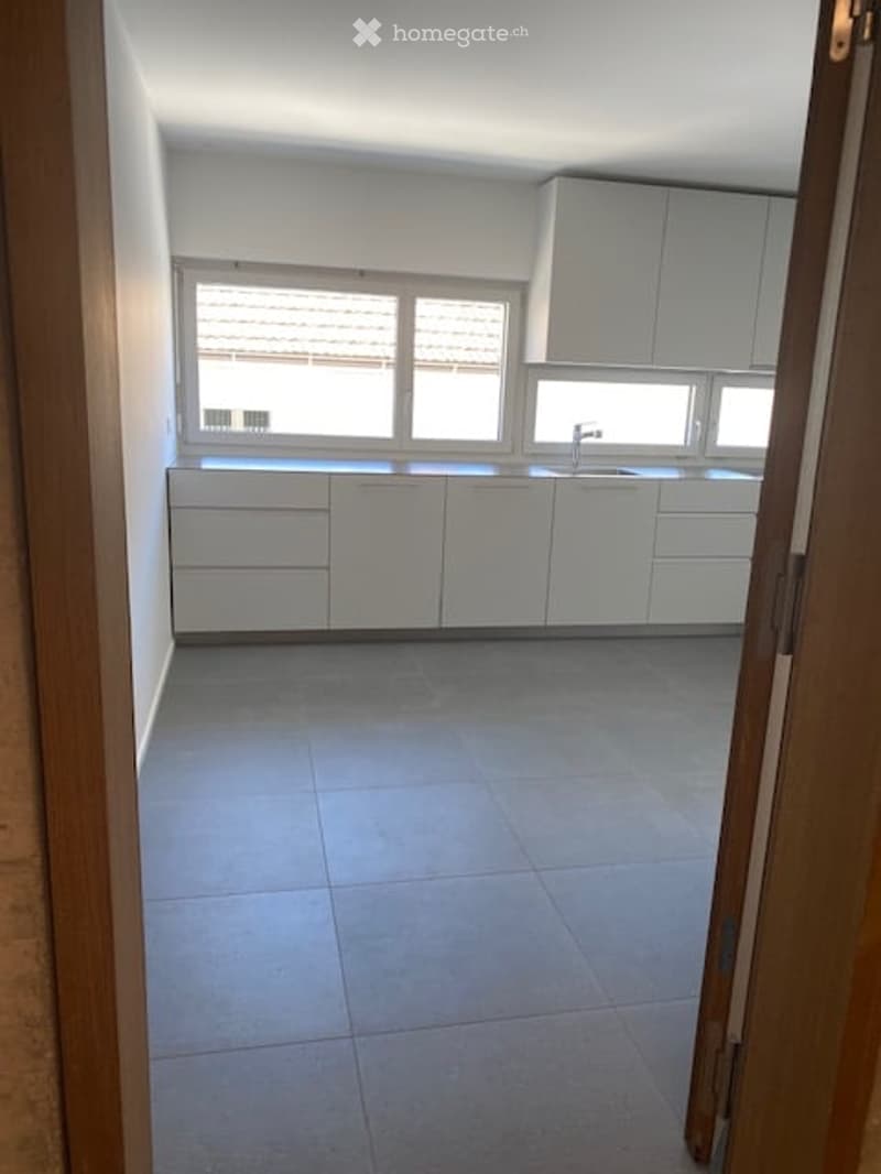 Modern, minimalist kitchen with white cabinets, a central island, and a large window providing natural light. The floor is tiled in a light grey color.