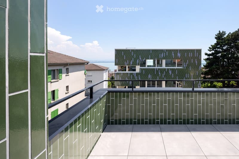 Modern balcony with a sleek black railing, tiled floor, and a view of a green building with a patterned facade and surrounding trees.