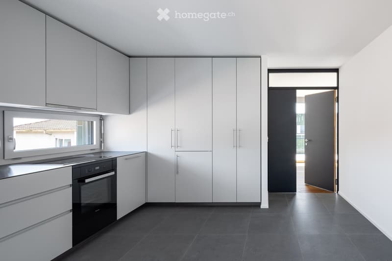 Modern kitchen with white cabinets, black oven, and dark tiled floor. Features a large window and an open doorway leading to another room.