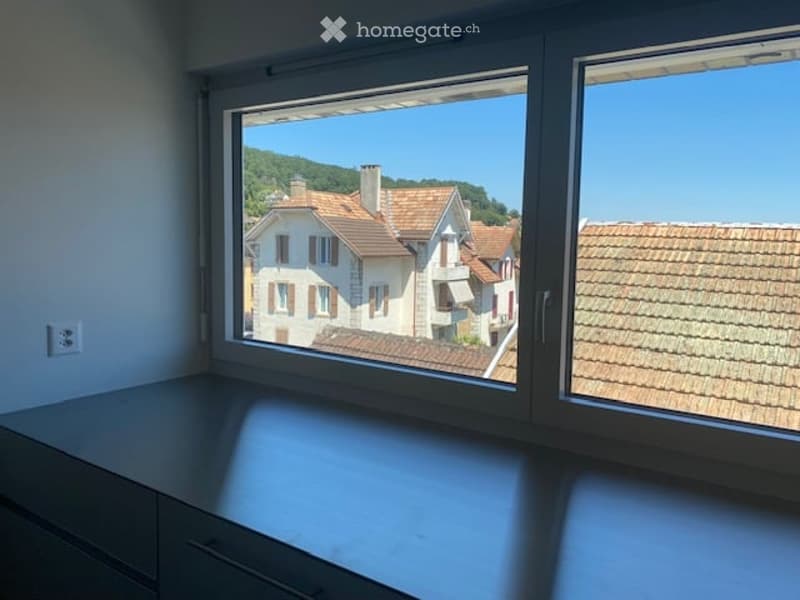 Empty room with large windows offering a view of a town with tiled roofs and surrounding hills. The room has a modern, minimalist design with a grey cabinet and white walls.