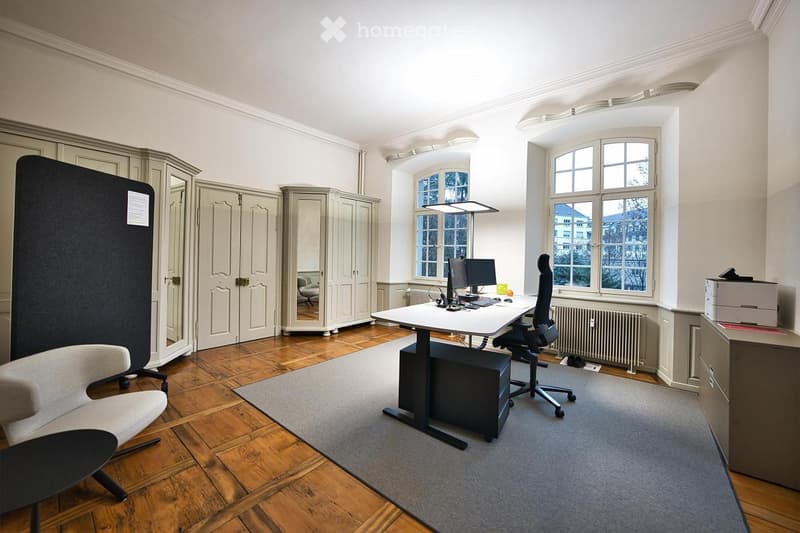 Office space with wooden floor, large windows, white table, black chair, grey cabinet, and printer.