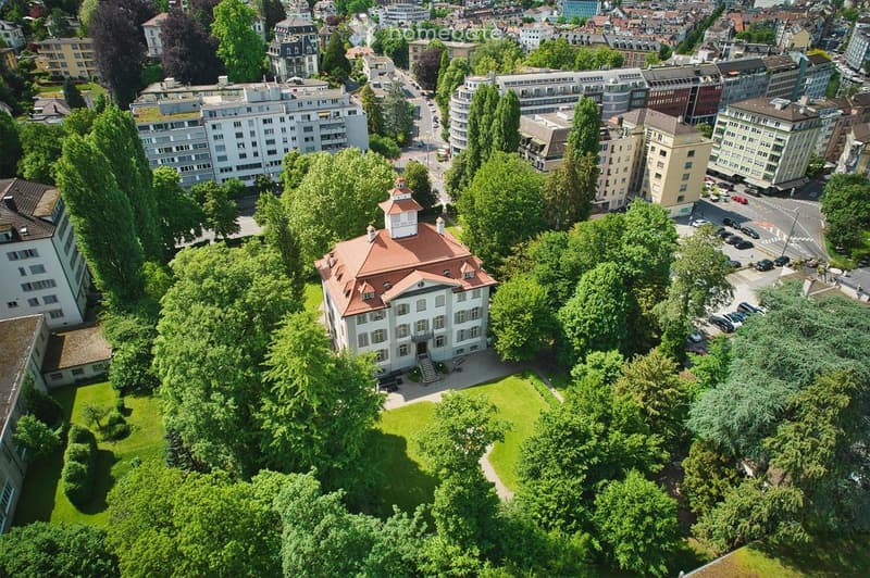 Aerial view of a large, classical-style building with a red-tiled roof and white facade, surrounded by lush green trees and a well-maintained lawn, situated in an urban area with other buildings and roads visible in the background.