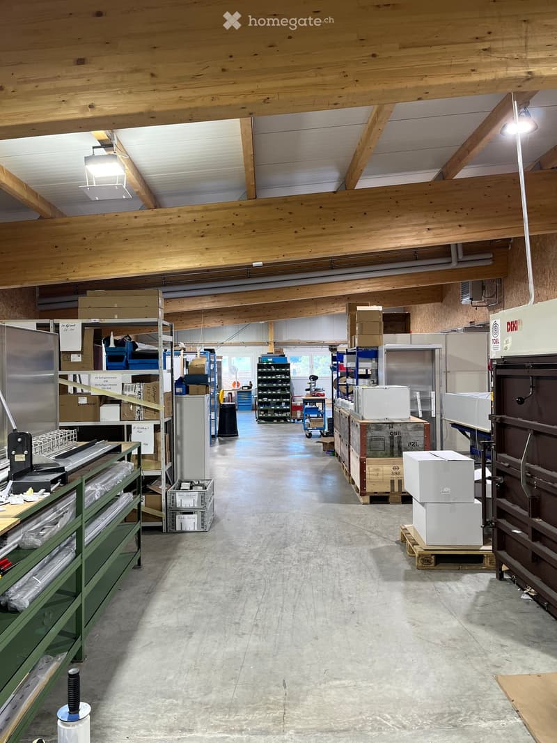 A spacious industrial or storage area with exposed wooden beams, concrete floor, and various storage equipment including shelves, refrigerators, and stacked cardboard boxes.