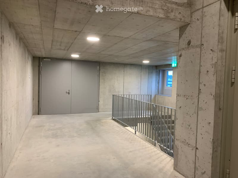 Concrete corridor with a staircase leading upwards, featuring a metal railing and a closed door at the end.