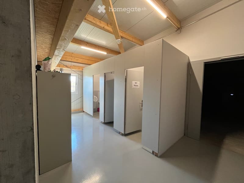 Empty hallway with white walls, grey floor, and several stall doors leading to bathrooms.
