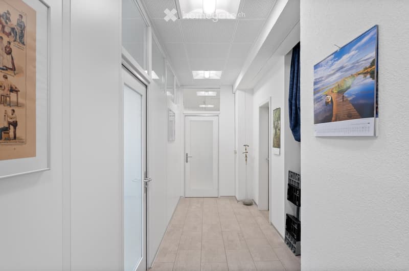 Modern, minimalist hallway with white walls, tiled floor, and large windows. Features artwork, a calendar, and a small black storage unit.