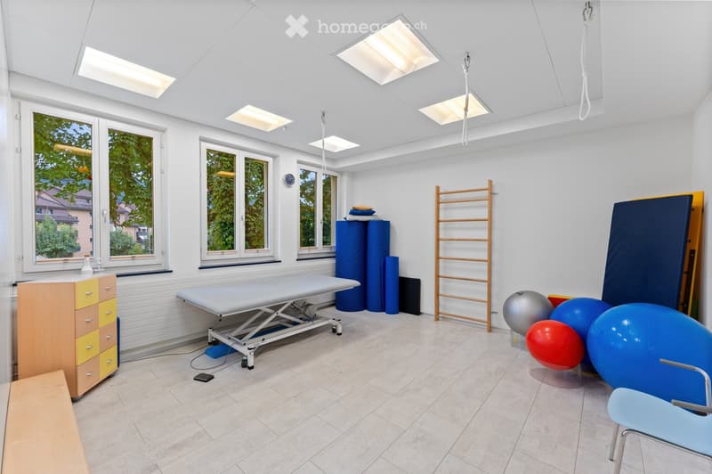 A well-lit therapy room with large windows, a treatment table, exercise balls, a ladder, and a drawer unit.