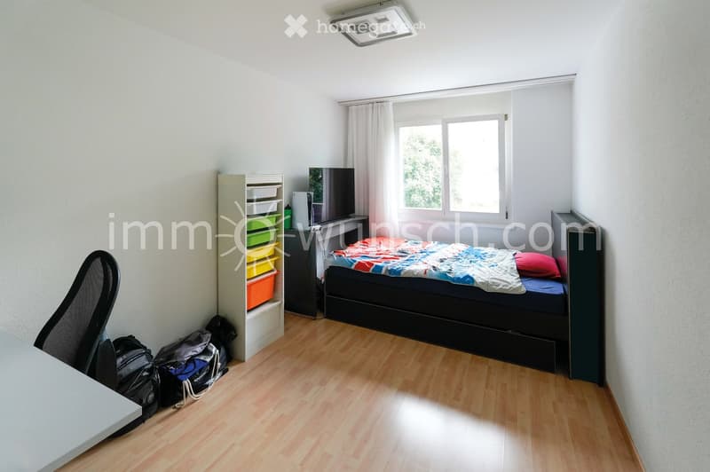 Bright bedroom with wooden flooring, large window with white curtains, bed with colorful bedding, chair, shelf with storage boxes, and a mirror on the wall.