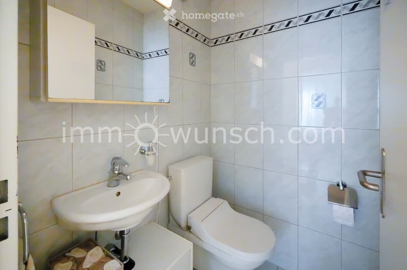 Modern bathroom with white tiles, a sink, toilet, mirror, and cabinet.
