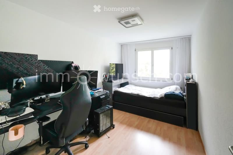 Modern bedroom with a large bed, desk with multiple screens, ergonomic chair, and a window with white curtains.