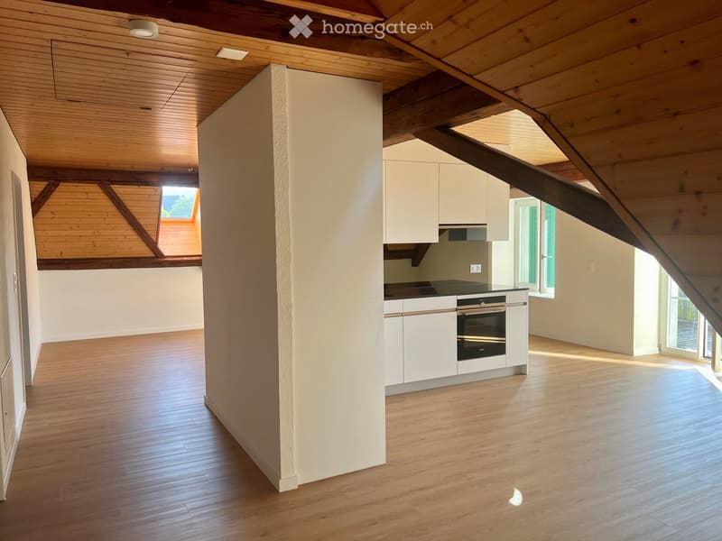 Open-plan attic space with wooden beams, light wood flooring, white cabinetry, and an integrated oven. Skylight and large windows provide natural light.