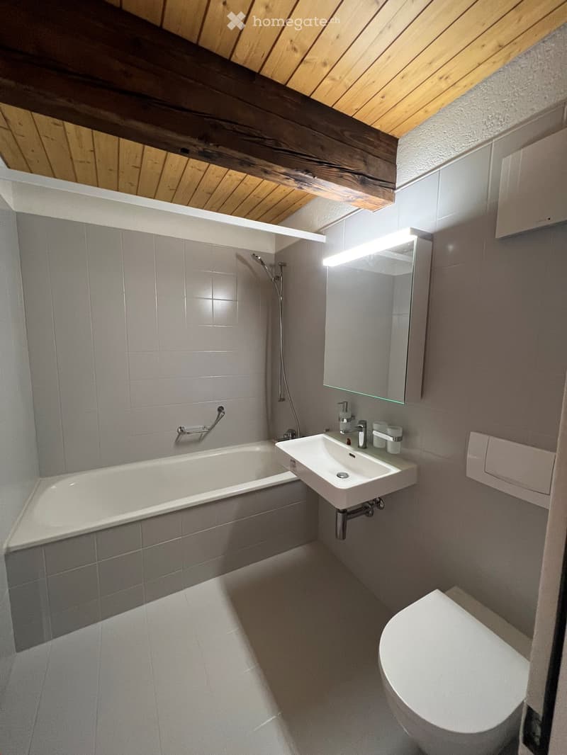 Modern bathroom with a bathtub, sink, toilet, mirror, and showerhead. The room features grey tiles and a wooden slanted ceiling.