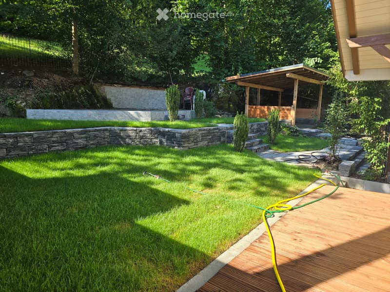 Well-maintained garden with a wooden deck, green lawn, stone walls, and a covered outdoor seating area.