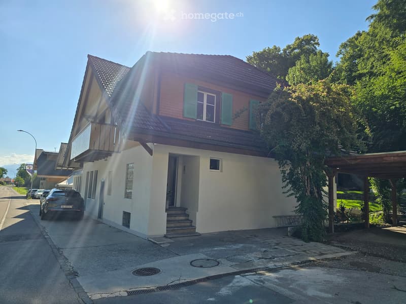 Two-story house with a red-tiled roof, white walls, and green shutters. Features a covered entrance, a small balcony, and a driveway with a parked car.