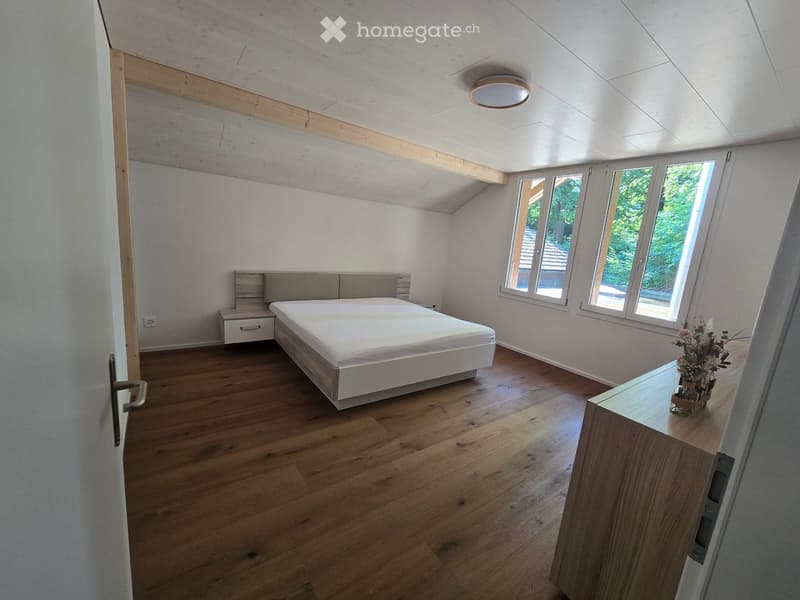 Modern bedroom with wooden flooring, large windows, a bed with a headboard, a nightstand, and a small table with a vase.