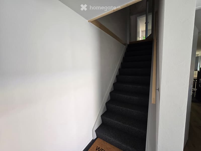 A modern staircase with dark carpeted steps and white walls, featuring a wooden handrail and a partially open door leading to another room.