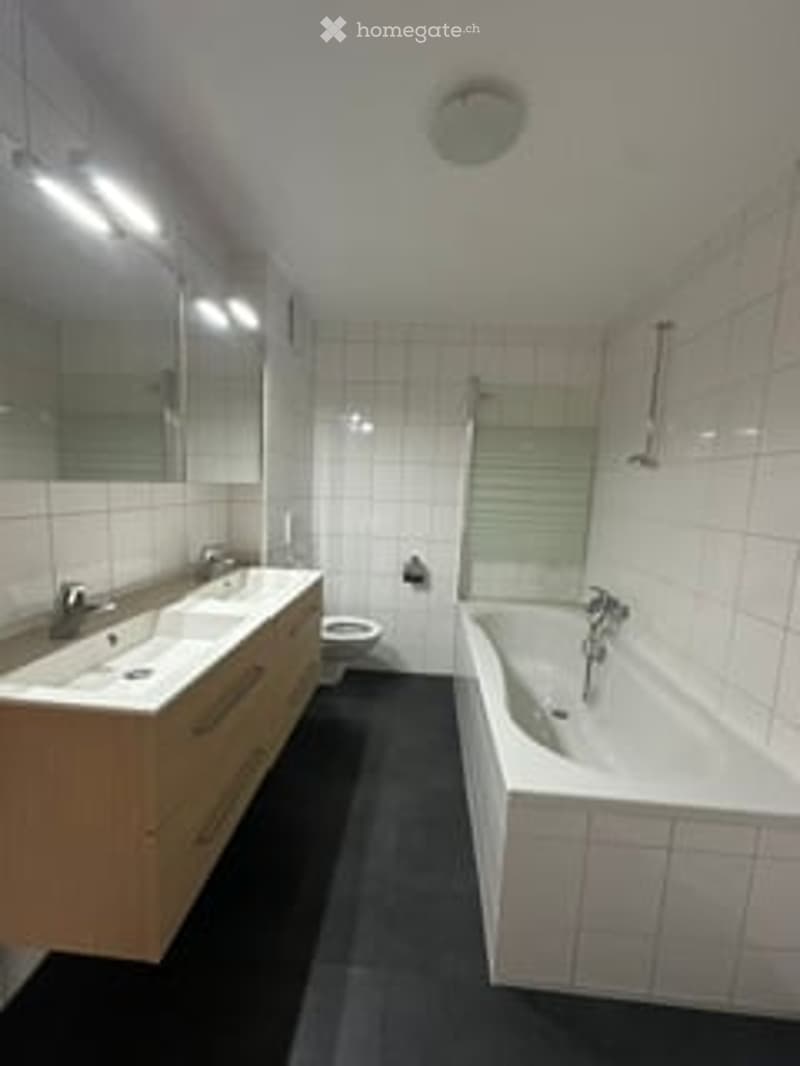 Modern bathroom with white tiles, a large bathtub, a sink with a mirror, and a toilet. The room features sleek, contemporary design elements and ample lighting.