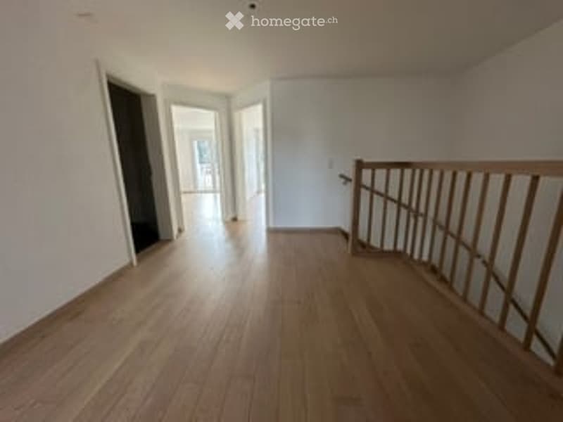 Empty room with wooden flooring and white walls, featuring a wooden staircase railing and open doorway leading to another room.