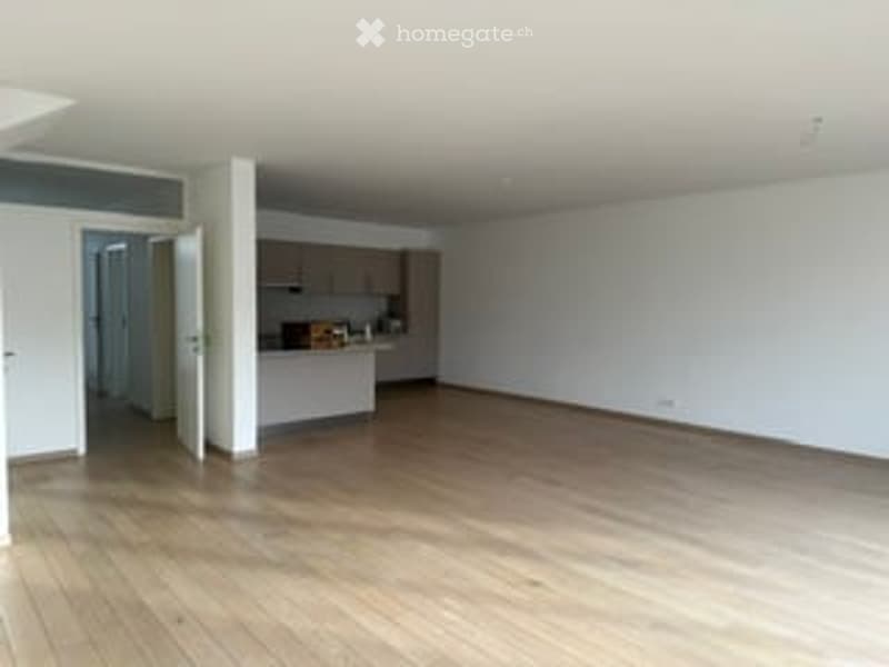 Empty room with wooden flooring, white walls, and a small kitchen area visible in the background.