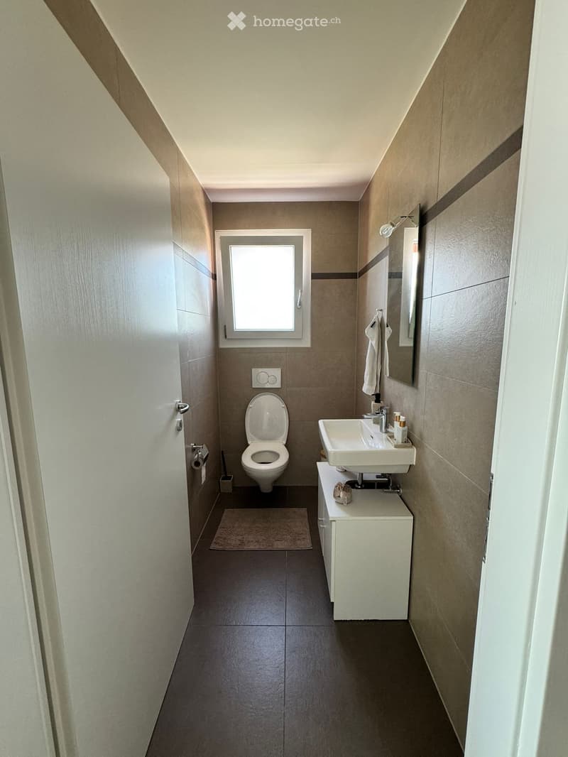 Modern bathroom with a toilet, sink, mirror, and towel. Features a window for natural light and a small rug on the floor.