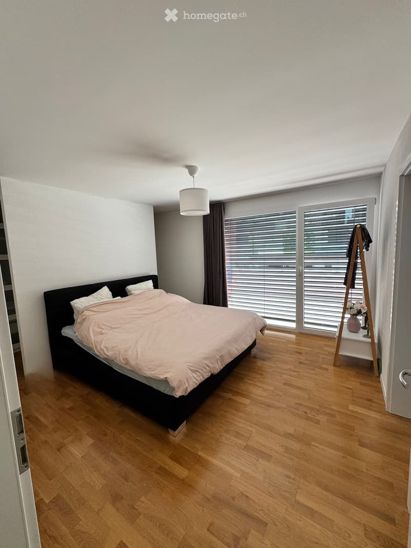 Modern bedroom with wooden flooring, large window with blinds, bed with beige bedding, pendant light, and coat rack with a vase on the shelf.