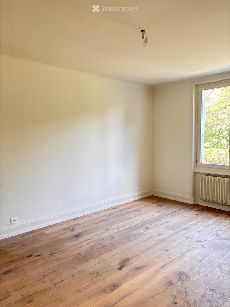 Empty room with light wooden flooring, white walls, and a window with a view of greenery. A radiator is visible under the window, and a single light fixture hangs from the ceiling.