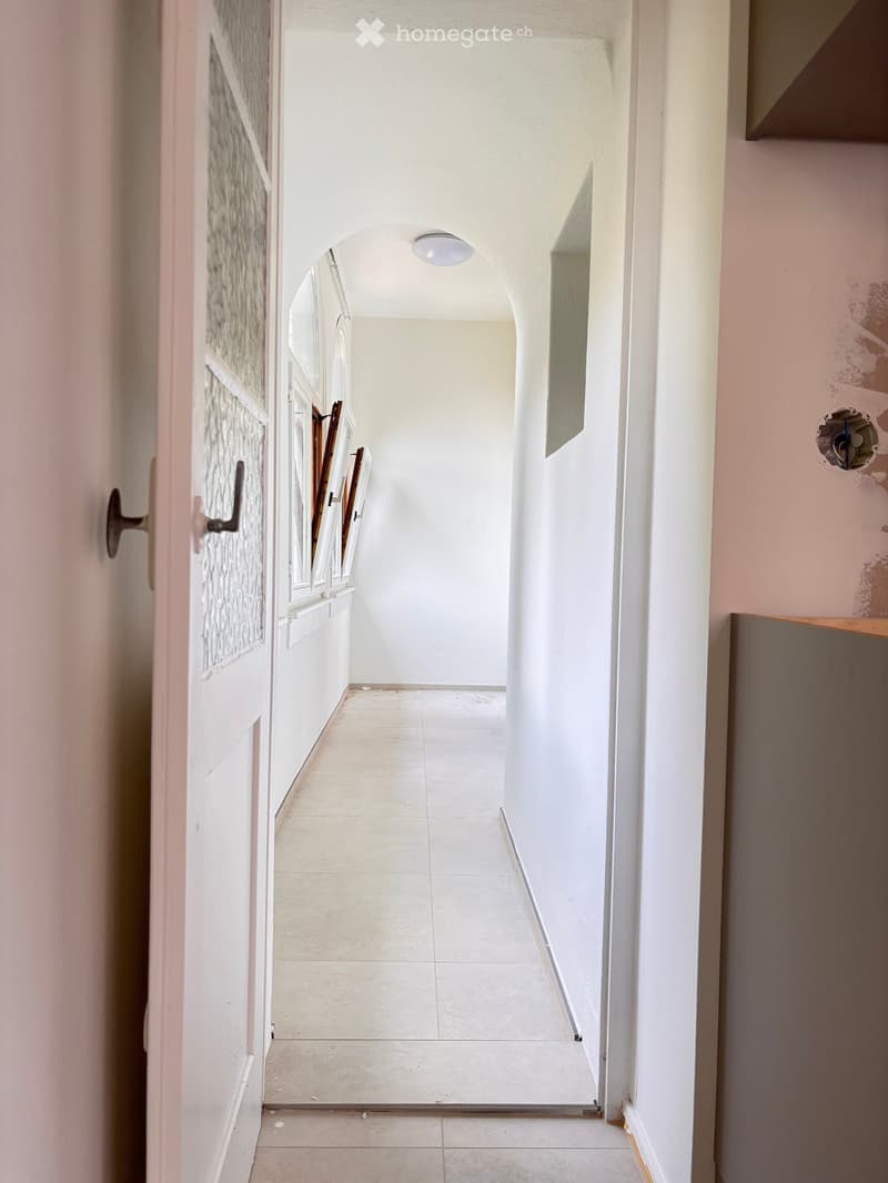 A narrow hallway with white tiled flooring, white walls, and a ceiling light. The hallway features an arched doorway at the end and a partially open door on the left side.