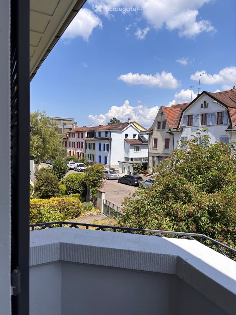 Balcony overlooking a residential street with houses, trees, and parked cars.