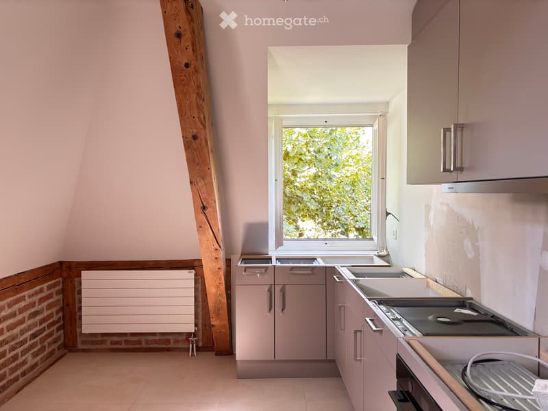 Modern kitchen with light pink cabinets, white countertop, and stainless steel appliances, featuring a large window with a view of greenery.