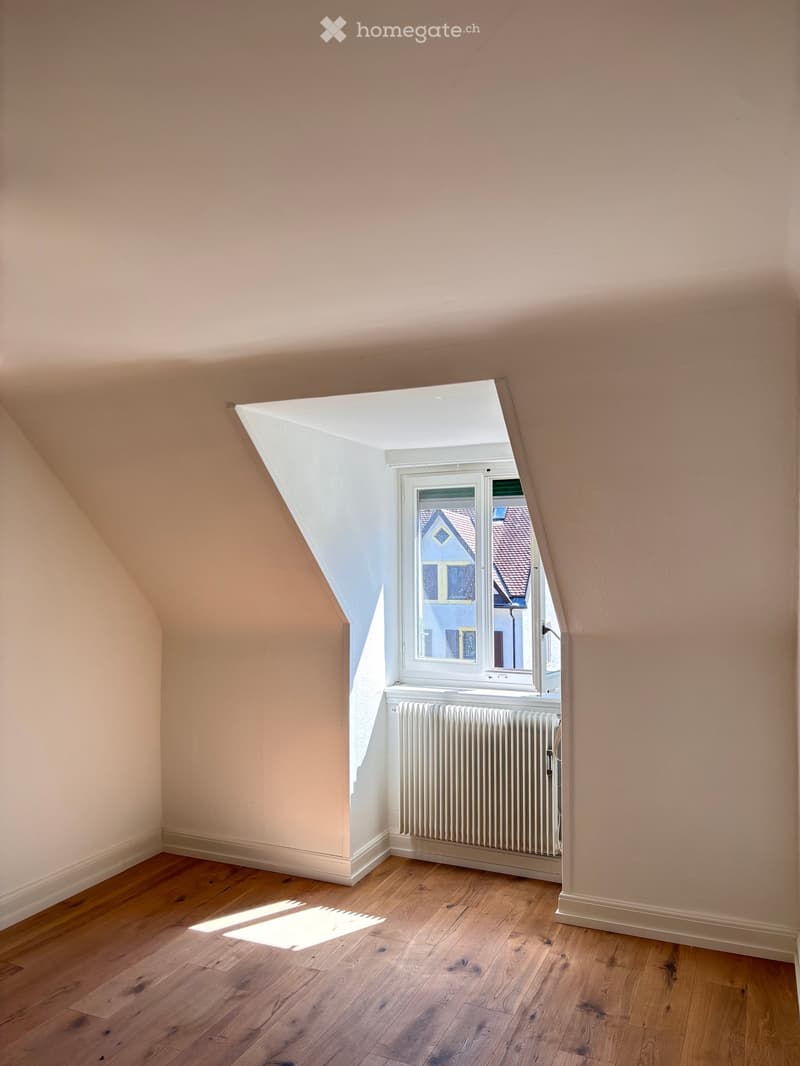 Empty attic room with wooden flooring, slanted ceilings, and a window showing a view of neighboring houses.