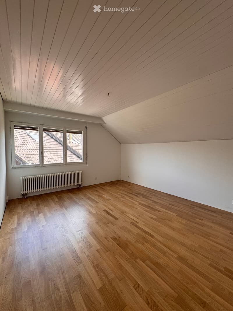 Empty attic room with wooden flooring, white walls, and a slanted wooden ceiling. Three windows offer a view of the roof.