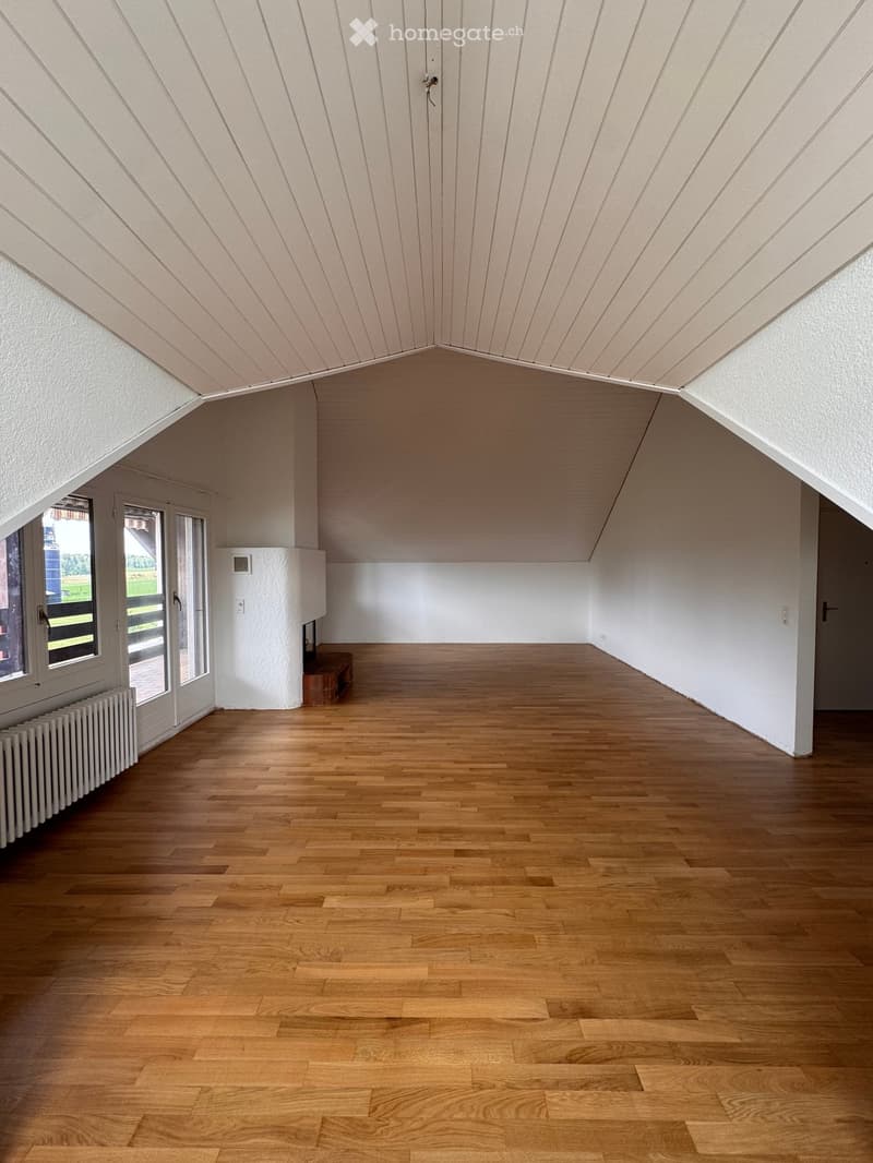 Empty room with wooden flooring, white walls, and a slanted ceiling. Features include a radiator, a small bench, and a window with a view of the outdoors.