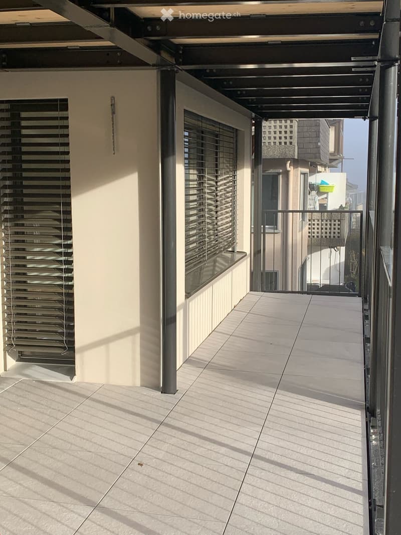 Modern balcony with tiled floor, metal railings, and large windows with blinds. The area is clean and well-lit with natural light.