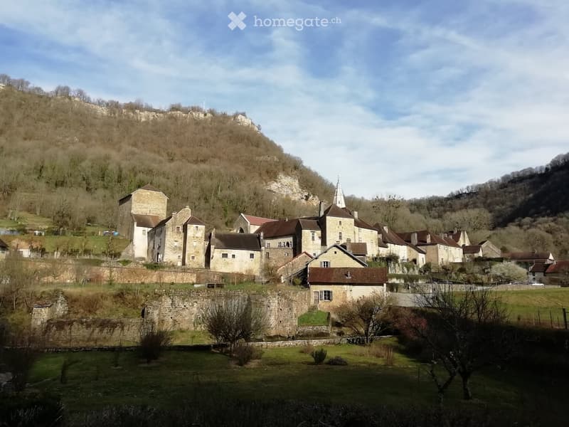 A cluster of old stone buildings with red-tiled roofs, surrounded by a stone wall and set against a hillside with sparse trees.