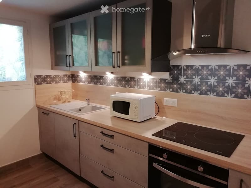 Modern kitchen with light wood cabinets, stainless steel appliances, and a decorative backsplash. Features include an induction stove, microwave, sink, and a bowl on the counter.