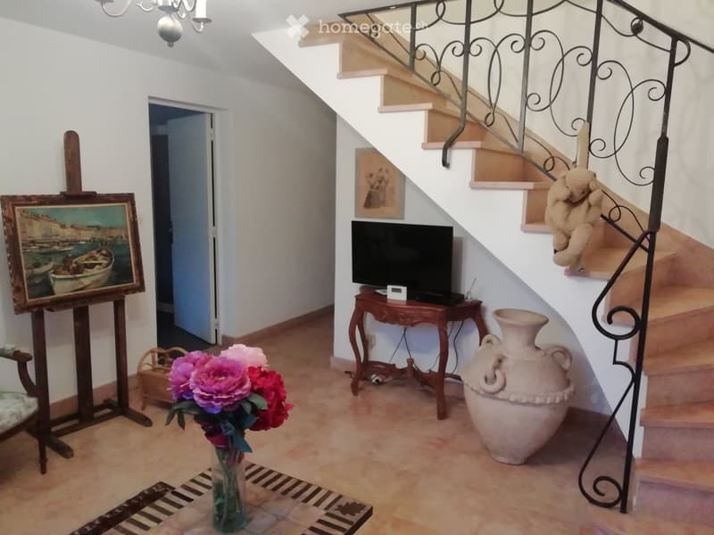 A well-lit hallway with a staircase, a small wooden table with a TV, a large ceramic vase, a painting on an easel, and a flower arrangement in a vase on the floor.