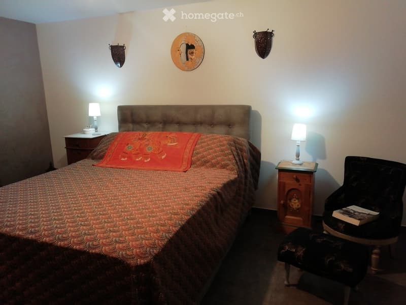 Cozy bedroom with a tufted headboard bed, patterned bedspread, nightstands with lamps, and a decorative chair with a book on it.