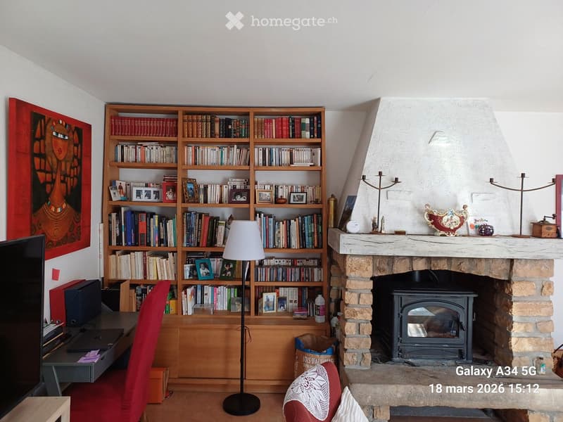A cozy living room with a large bookshelf filled with books and framed photos, a fireplace with decorative items on the mantel, a red armchair, a floor lamp, and a small table with a laptop.