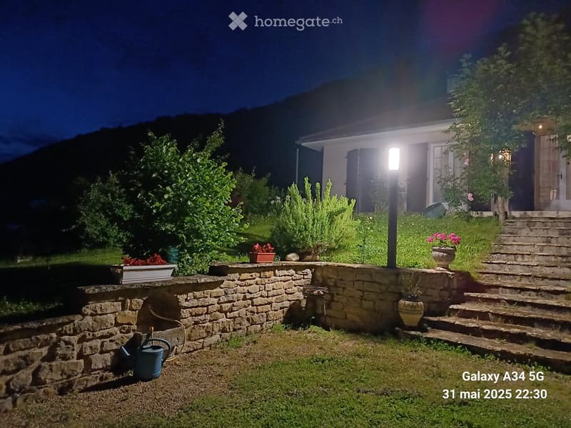 Nighttime garden with stone wall, potted plants, steps leading to a house, and outdoor lighting.