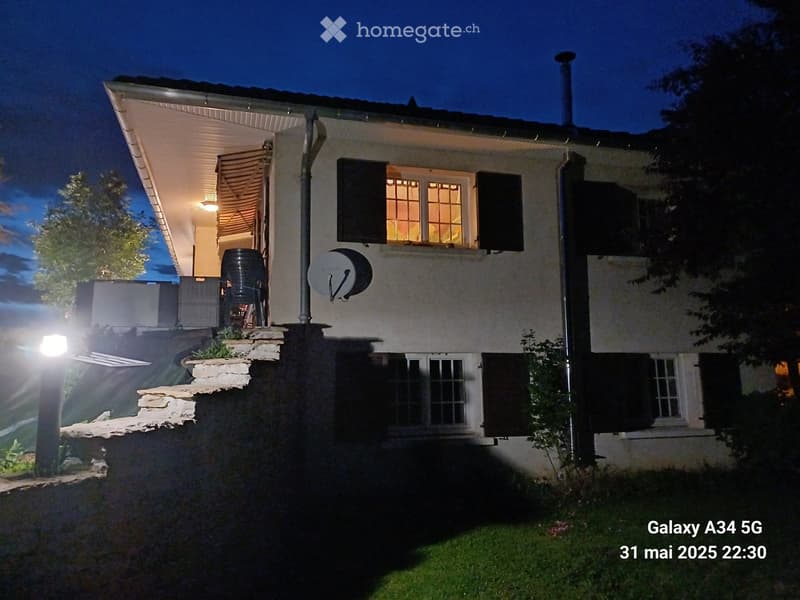 Two-story house with white exterior, multiple windows, and a covered entrance area. The house features a raised ground floor and is surrounded by a well-maintained lawn.
