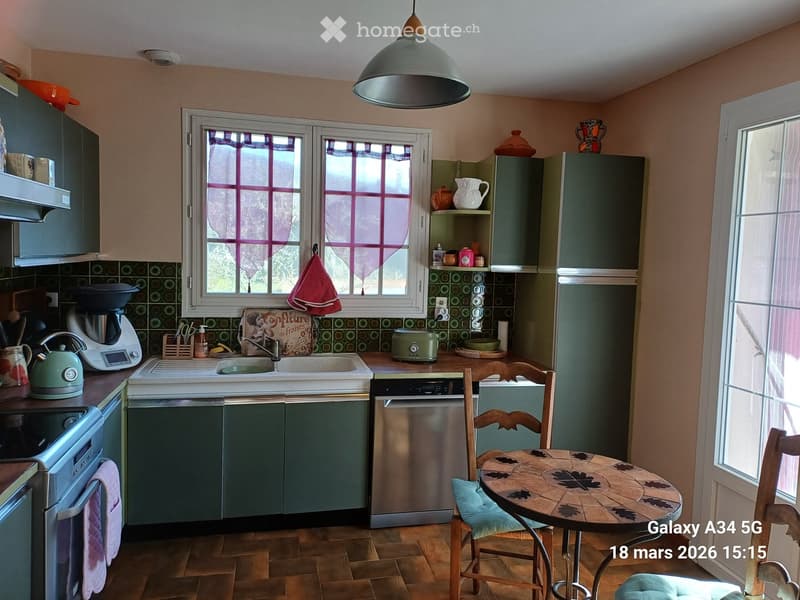A cozy kitchen with green cabinets, a stainless steel dishwasher, a coffee maker, a toaster, a teapot, and a small dining area with a round table and chairs.