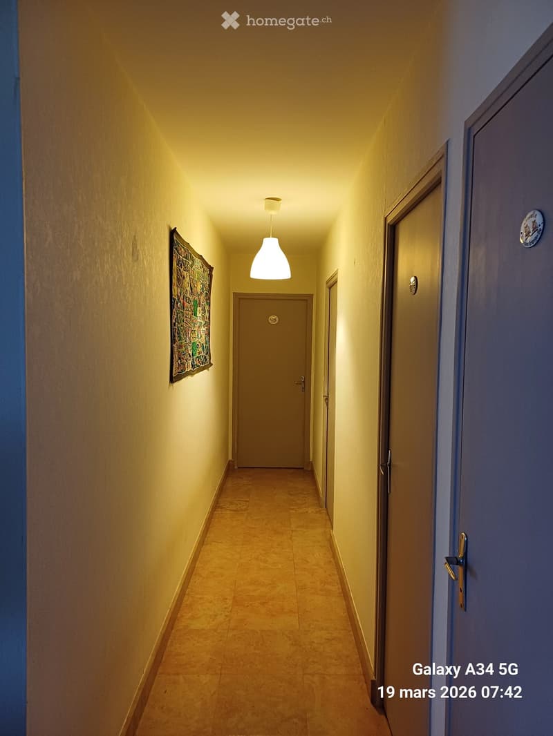 A narrow hallway with beige tiled flooring, light yellow walls, and a single hanging light fixture. Doors are visible on both sides, and a decorative piece of art hangs on the left wall.