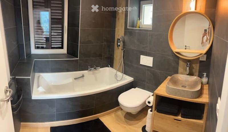 Modern bathroom with dark grey tiles, a bathtub, toilet, sink, and a round mirror above the sink. Wooden shelf holds towels and toiletries.