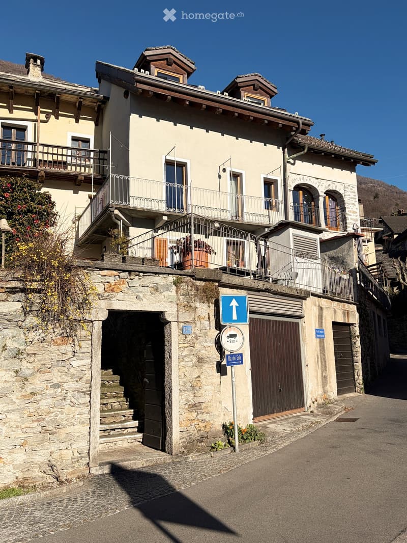 The building has multiple stories, exposed stonework on the lower portion, balconies with iron railings, stairs leading to a lower entrance, two garages with brown doors, and a blue sign indicating height restrictions.