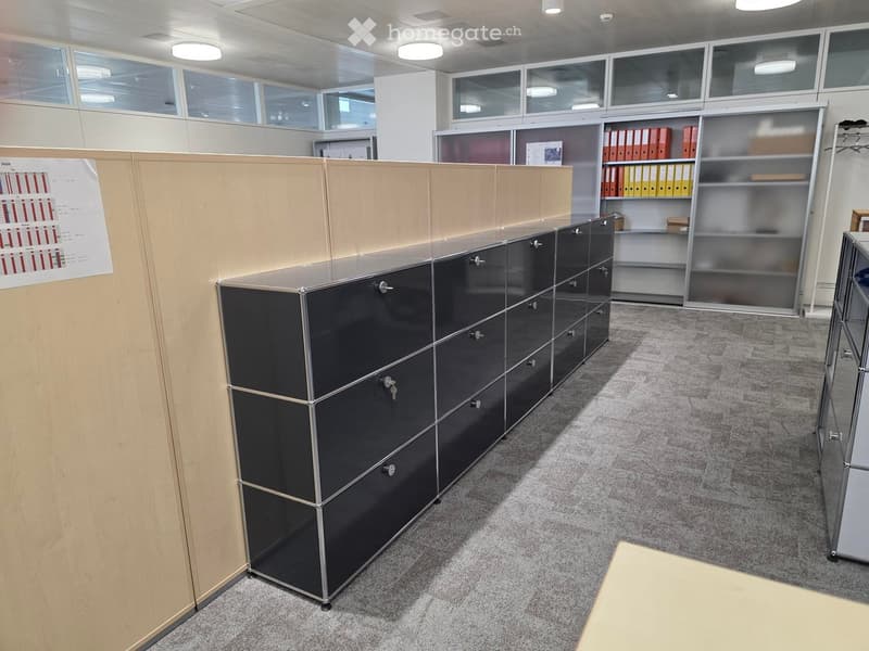 Modern office space with grey carpet, glass partitions, black metal cabinets, and shelves holding binders.